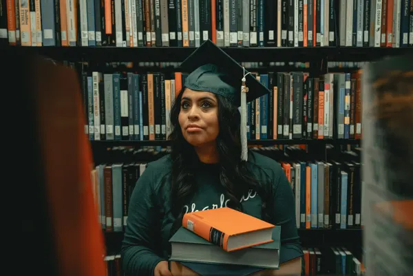 A graduate in a mortarboard holding a stack of books in a library.