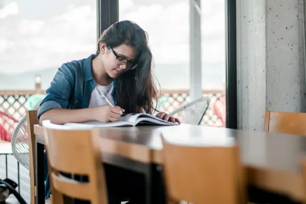A student writing in a notebook at a library table.