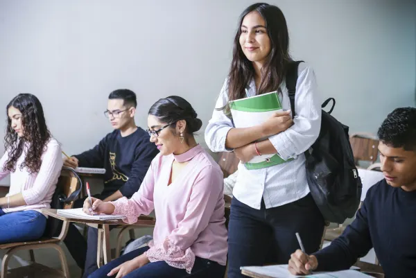 Students working at their desks in a bright classroom, one arriving with her textbooks.